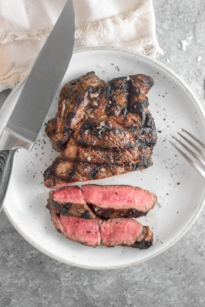 Ribeye Steak with criss cross grill marks cooked to medium rare pink inside on a white plate with grey background and silver fork on the right
