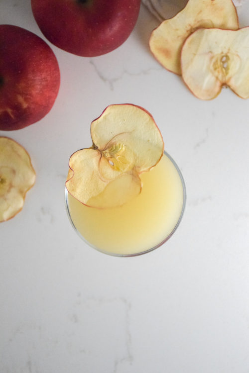 Small cocktail glass with filled with golden cocktail and garnished with dried apple slice on a white background with red apple in background