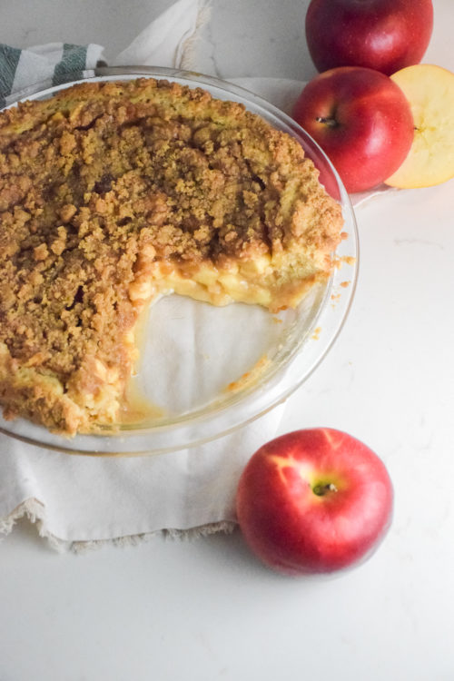 Apple Pie with crumble topping and cookie crust in a glass pie plate with red apples in background