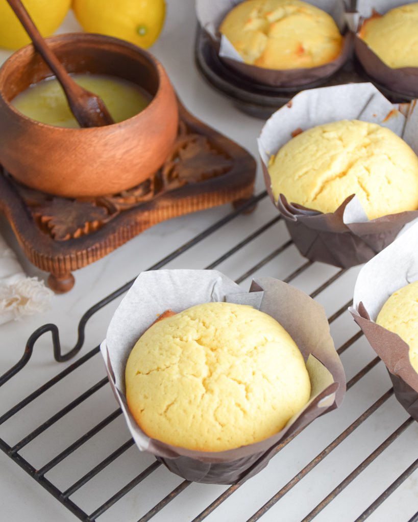 yellow lemon cakes in paper muffin wrappers on metal cooling rack with brown wooden bowl with lemon glaze in it