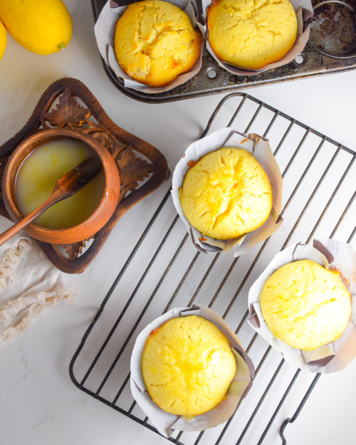 yellow lemon cakes in paper muffin wrappers on metal cooling rack with brown wooden bowl with lemon glaze in it