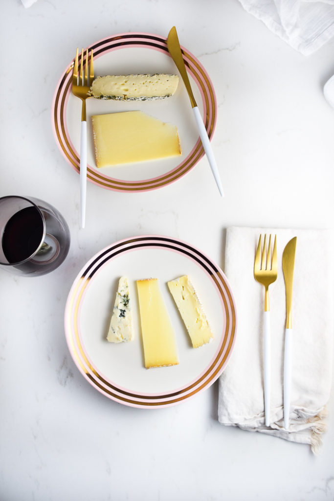 Three pieces of french cheese on a plate with a fork and knife on the right and a glass of red wine on the left 