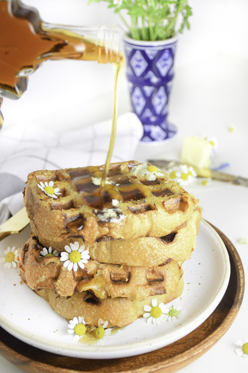 syrup drizzling on a stack of french toast waffles on a white plate with chamomile flowers and blue and flower vase in background