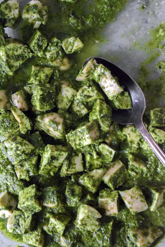 Bite sized cubes of chicken breast covered in a green pesto sauce in a clear baking dish with a spoon