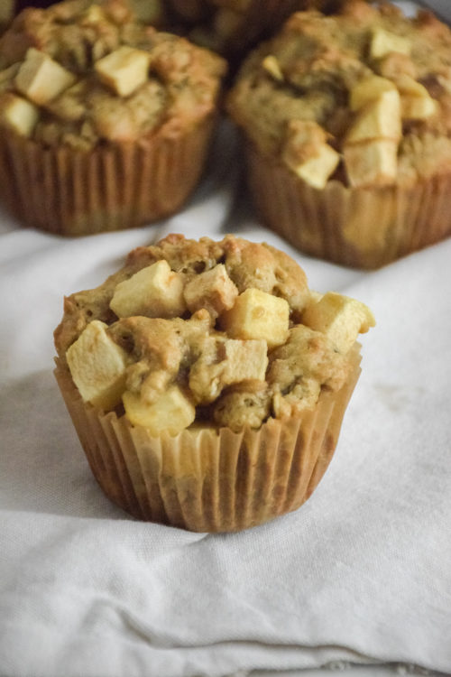 Muffin with chunks of apple on a white background with red apple and muffins in the background
