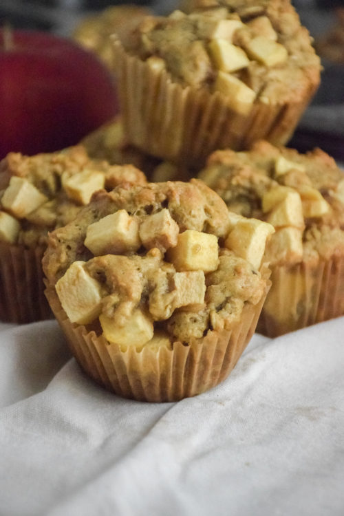 Muffin with chunks of apple on a white background with red apple and muffins in the background