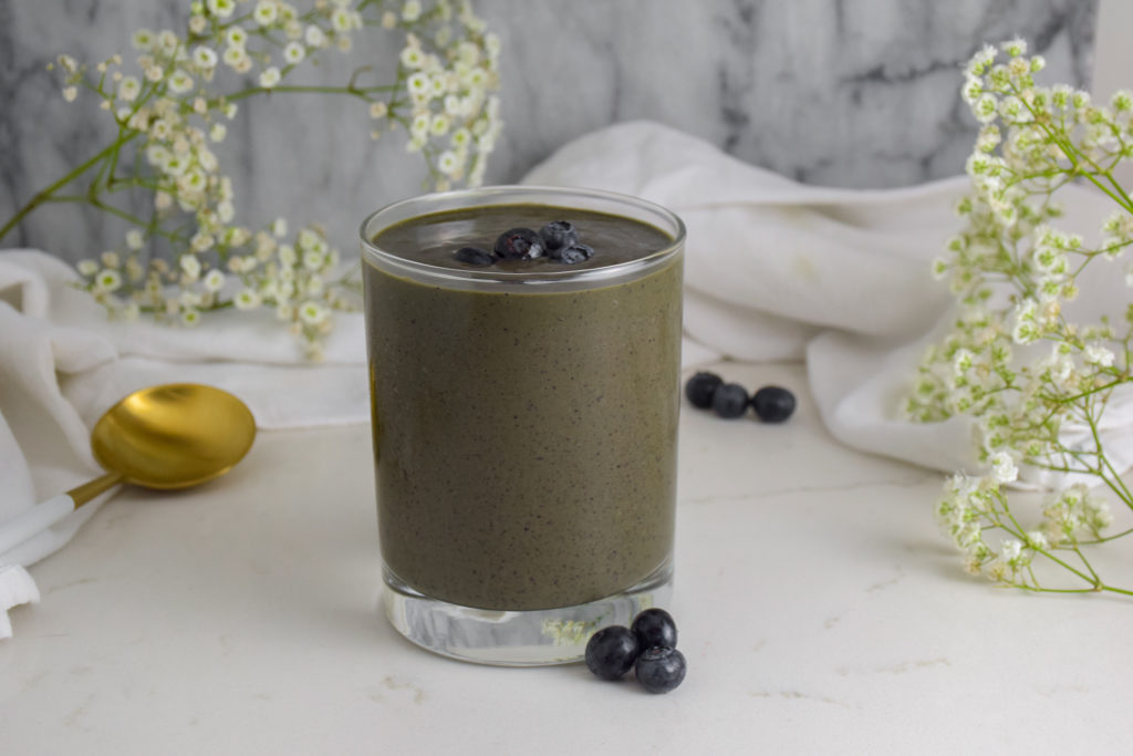 Blue smoothie in a clear glass with fresh blueberries on top. Marble background with flowers