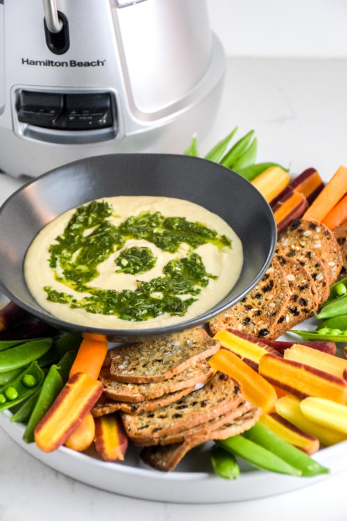 creamy white bean dip with herb oil swirled on top in a black bowl with vegetables and crackers on the side