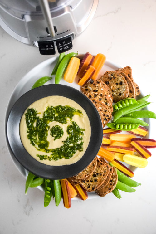 creamy white bean dip with herb oil swirled on top in a black bowl with vegetables and crackers on the side