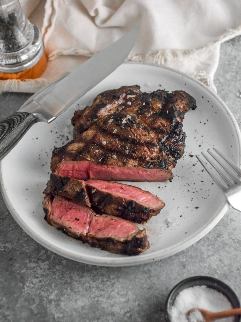 Ribeye Steak with criss cross grill marks cooked to medium rare pink inside on a white plate with grey background and silver fork on the right 