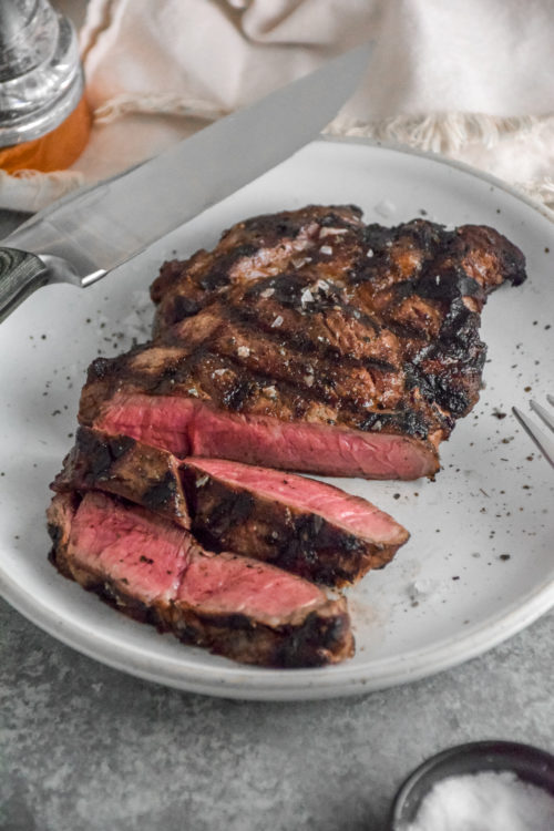 Ribeye Steak with criss cross grill marks cooked to medium rare pink inside on a white plate with grey background and silver fork on the right