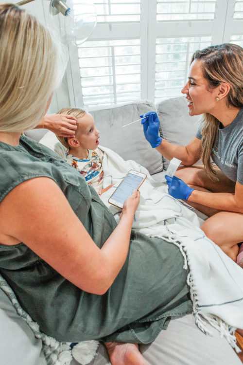 child sitting next to mother on couch with a nurse with blue medical gloves giving throat swab test