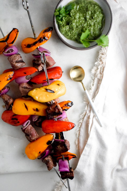 Sirloin Steak with red and orange baby bell peppers and red onion on metal skewers with black grill marks on a white background