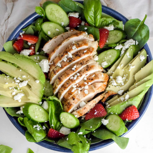 Salad with sliced grilled chicken breast in the middle and avocado and strawberries and feta cheese on top all on a white background