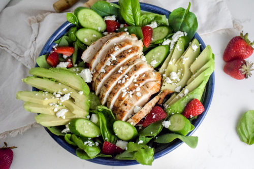 Salad with sliced grilled chicken breast in the middle and avocado and strawberries and feta cheese on top all on a white background