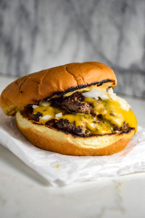 Burger with two thin patties on a bun with cheese and onions on a crumbled white paper on the grey marble background