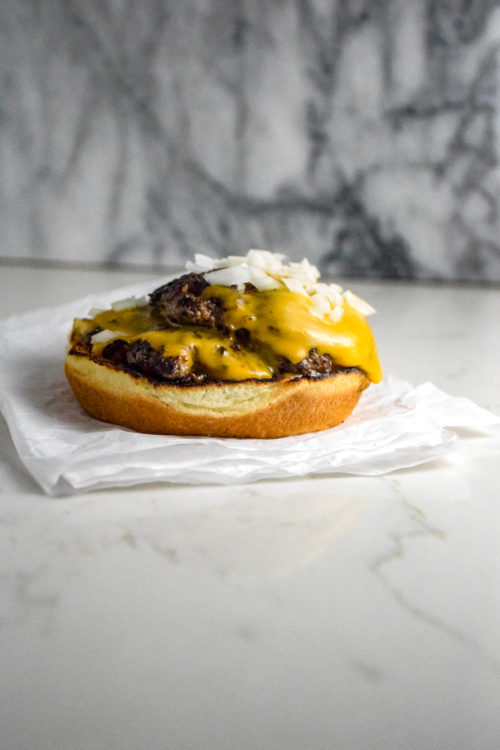 Burger with two thin patties on a bun with cheese and onions on a crumbled white paper on the grey marble background
